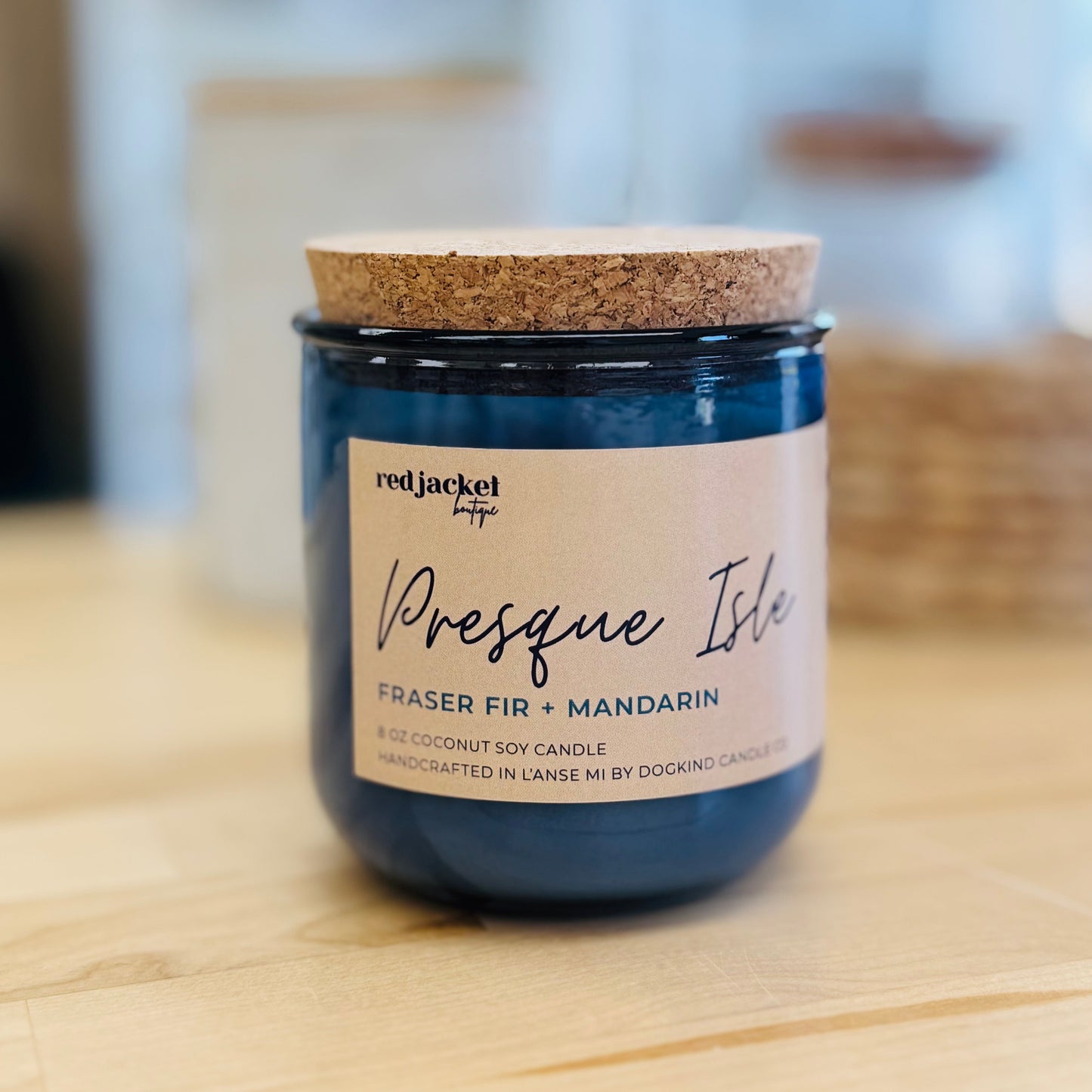 
                  
                    A blue glass jar candle with a cork lid and a cream label reading 'redjacket' and 'Presque Isle', placed on a light wood surface with a blurred indoor background.
                  
                