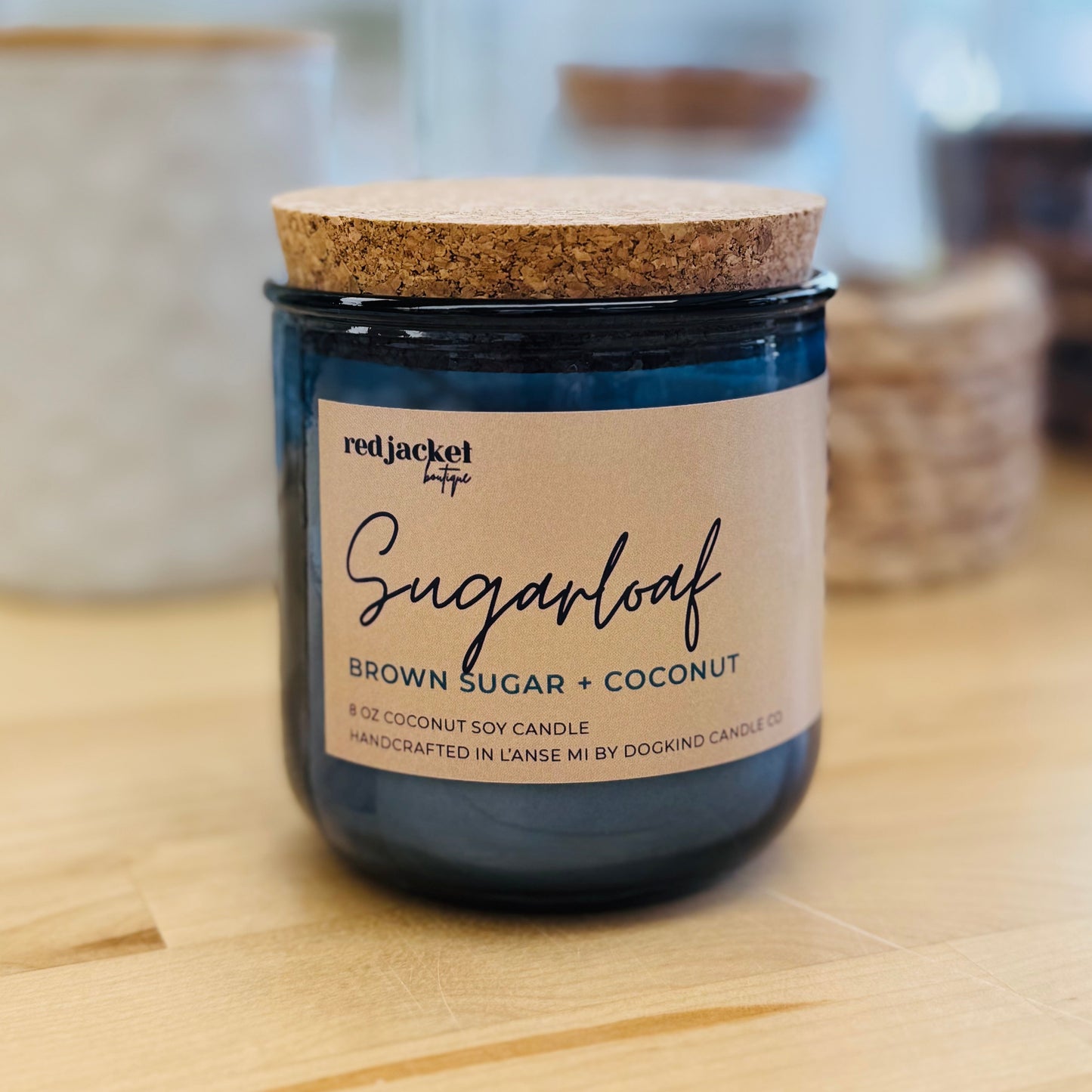 
                  
                    A jar candle labeled 'Sugarloaf' by redJacket Co, with brown sugar and coconut scent, featuring a dark blue glass jar, a cork lid, and a cream paper label, set on a light wood surface with a blurred background.
                  
                