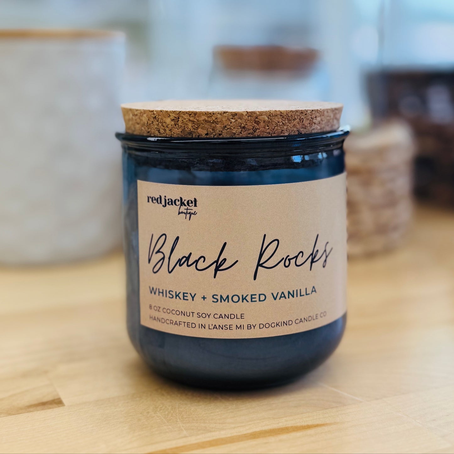 
                  
                    A dark blue glass jar candle with a cork lid and a beige label reading 'Black Rocks Whiskey + Smoked Vanilla' by Red Jacket, placed on a light wood surface with a softly blurred indoor background.
                  
                