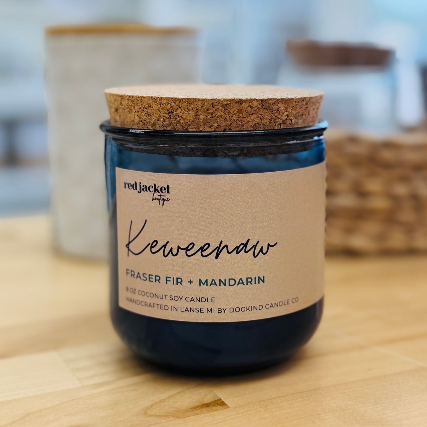 A dark blue glass jar candle with a natural cork lid and a cream paper label reading 'Kewenaw' and 'Red Jacket Candle Co.' on a light wood surface with a softly blurred indoor background.