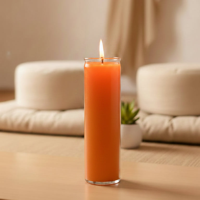 
                  
                    A tall orange pillar candle in a clear glass holder, lit with a small flame, standing on a light wood table in a softly blurred indoor setting with cushions and a small plant in the background.
                  
                