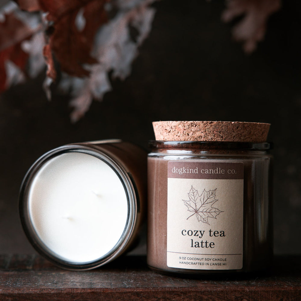 Two fall scented candles, one open and one with a cork lid, with 'cozy tea latte' label in front of an autumn oak leaves background.
