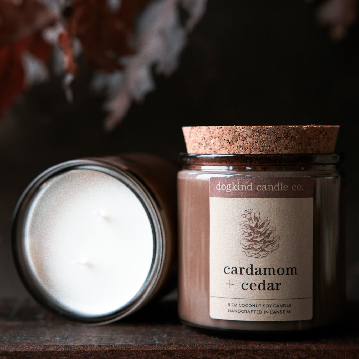 Two fall scented candles, one open and one with a cork lid, with a label reading 'cardamom + cedar' on a dark background in front of autumn oak leaves.