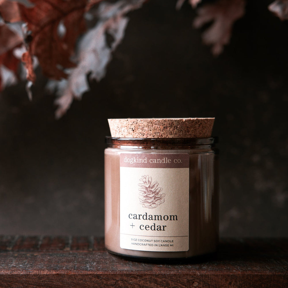 
                  
                    Fall scented Candle jar labeled 'cardamom + cedar' on a dark surface with a blurred natural background of autumn oak leaves.
                  
                
