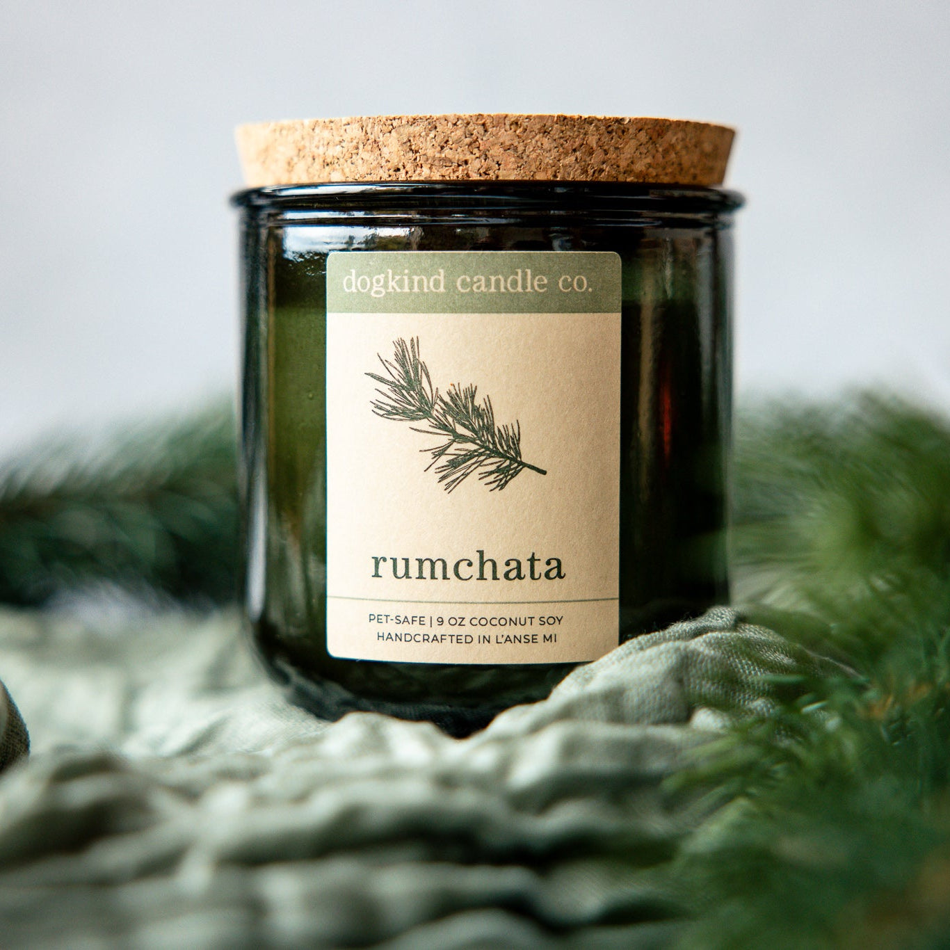 RumChata holiday scented candle from Dogkind Candle Co. on a textured surface with pine greenery in the background.