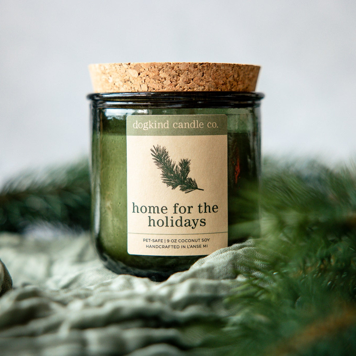 Home for the Holidays scented candle from Dogkind Candle Co. on a textured surface with pine greenery in the background.