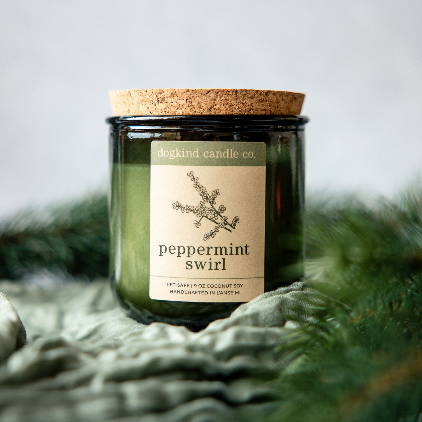 Peppermint Swirl holiday scented candle from Dogkind Candle Co. on a textured surface with pine greenery in the background.