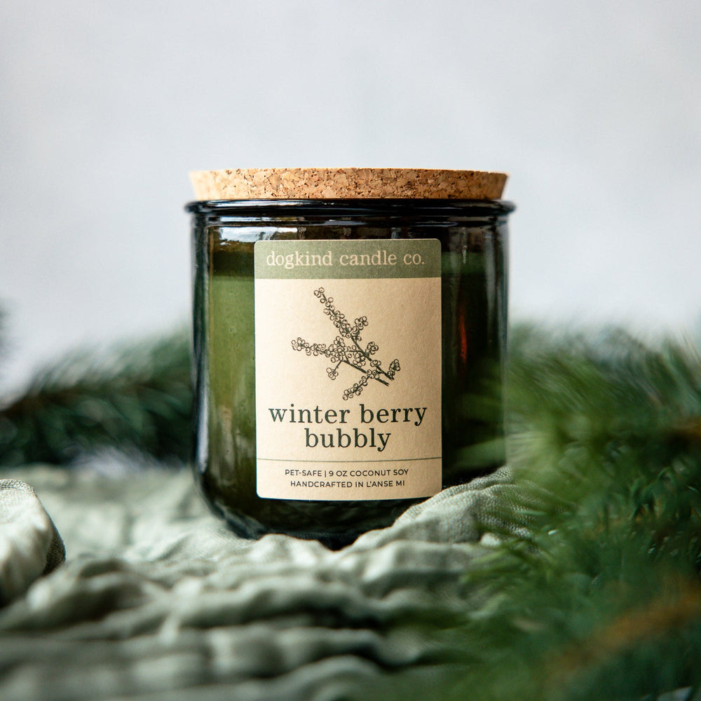 Winter Berry Bubbly holiday scented candle from Dogkind Candle Co. on a textured surface with pine greenery in the background.