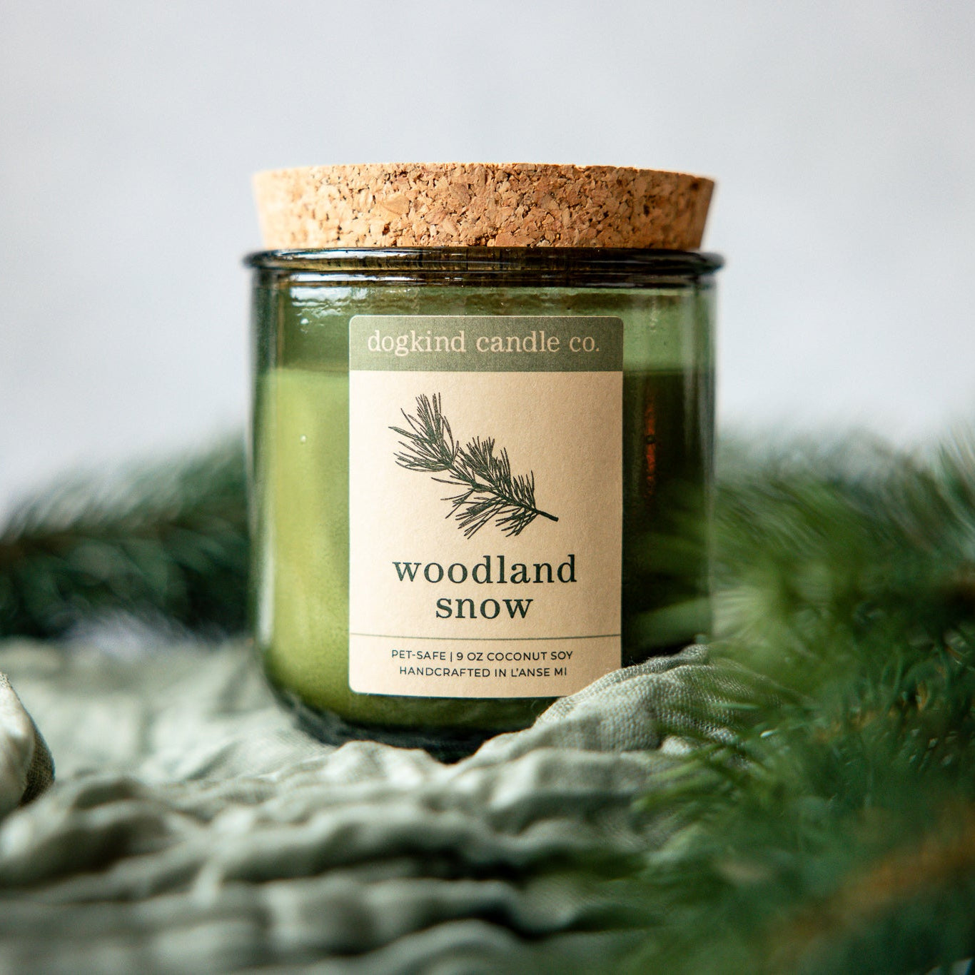 Woodland Snow holiday scented candle from Dogkind Candle Co. on a textured surface with pine greenery in the background.