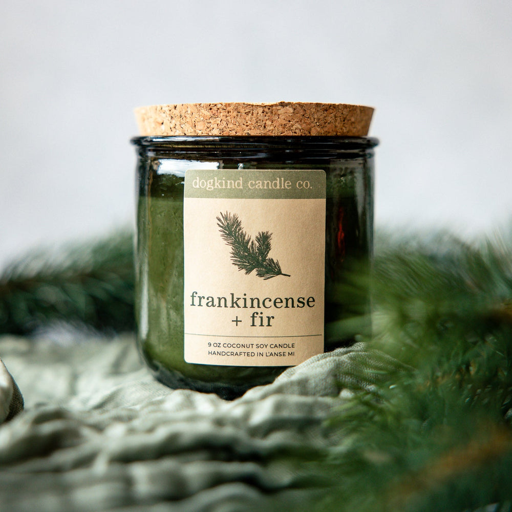 Frankincense + Fir  holiday candle from Dogkind Candle Co. on a textured surface with pine greenery in the background.