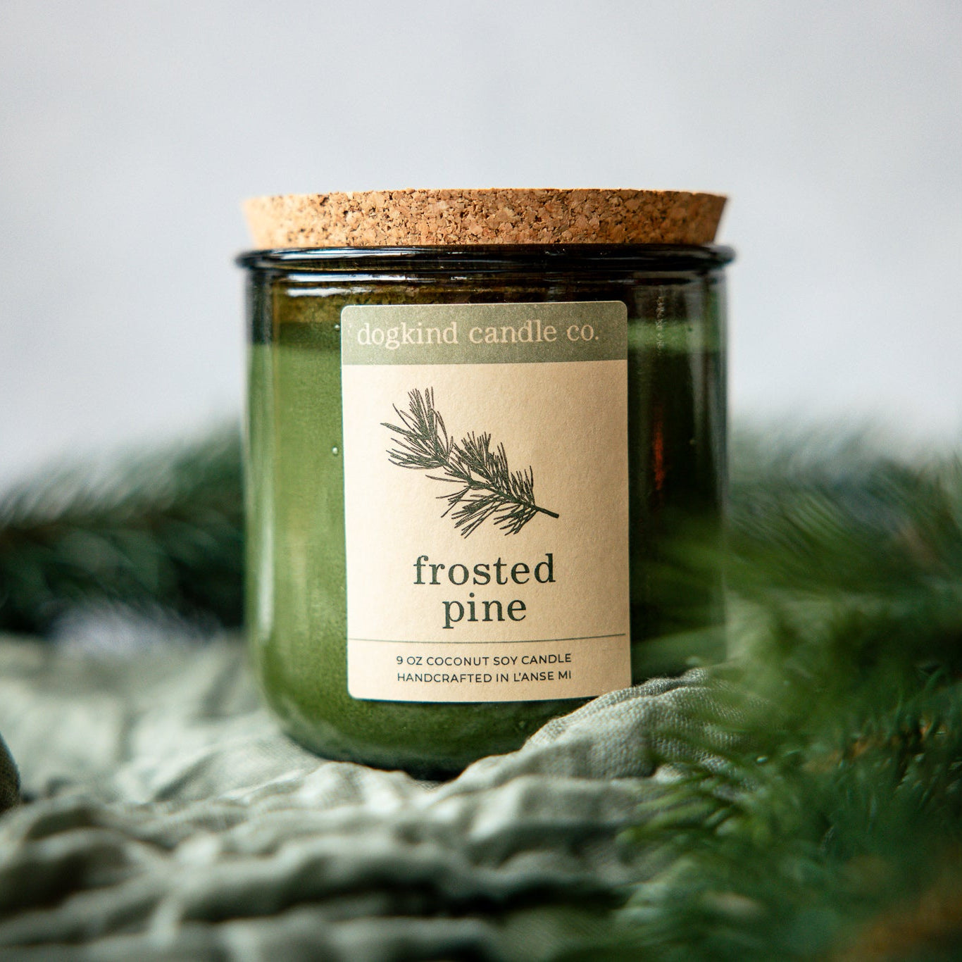 Frosted Pine holiday candle from Dogkind Candle Co. on a textured surface with pine greenery in the background.