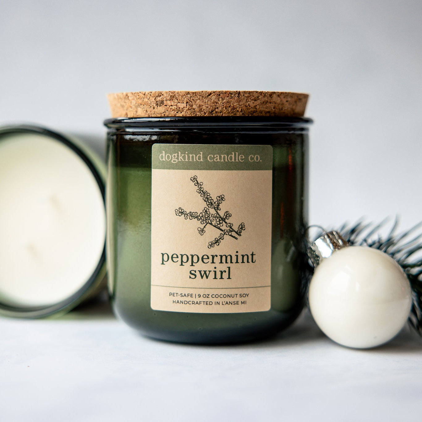 
                  
                    Peppermint Swirl holiday scented candle from Dogkind Candle Co. on a textured surface with pine greenery and Christmas ornament in the background.
                  
                