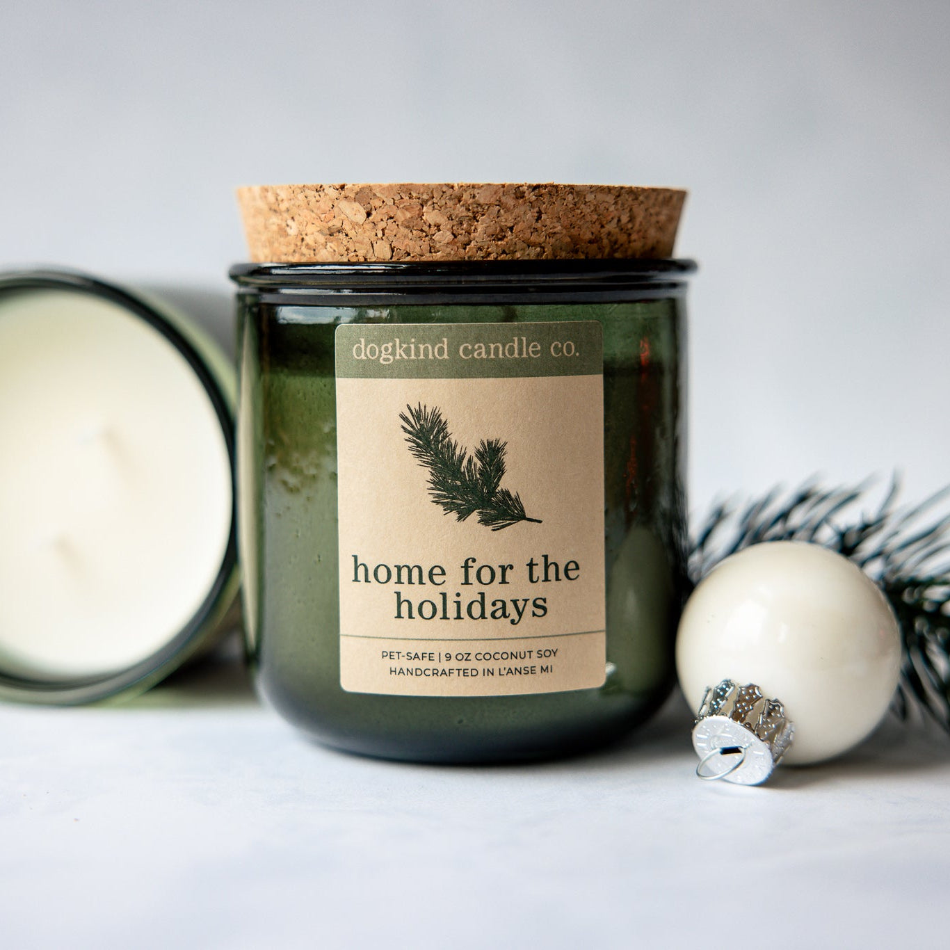Home for the Holidays scented candle from Dogkind Candle Co. on a textured surface with pine greenery and Christmas ornament in the background.