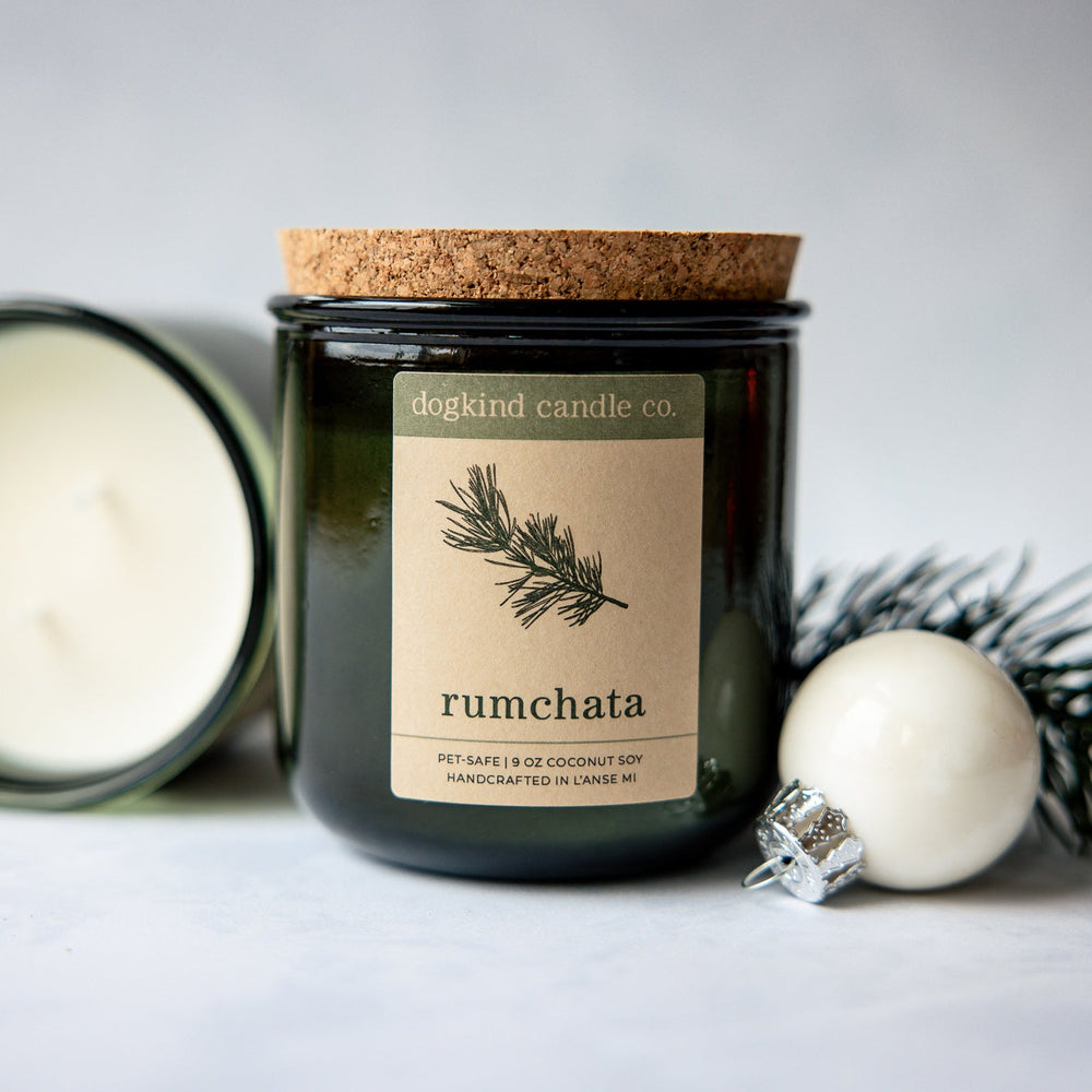 RumChata holiday scented candle from Dogkind Candle Co. on a textured surface with pine greenery and Christmas ornament in the background.