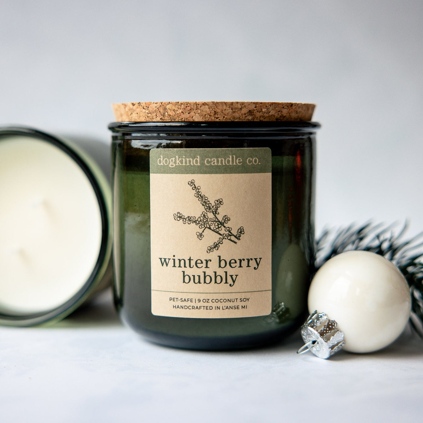 Winter Berry Bubbly holiday scented candle from Dogkind Candle Co. on a textured surface with pine greenery and Christmas ornament in the background.