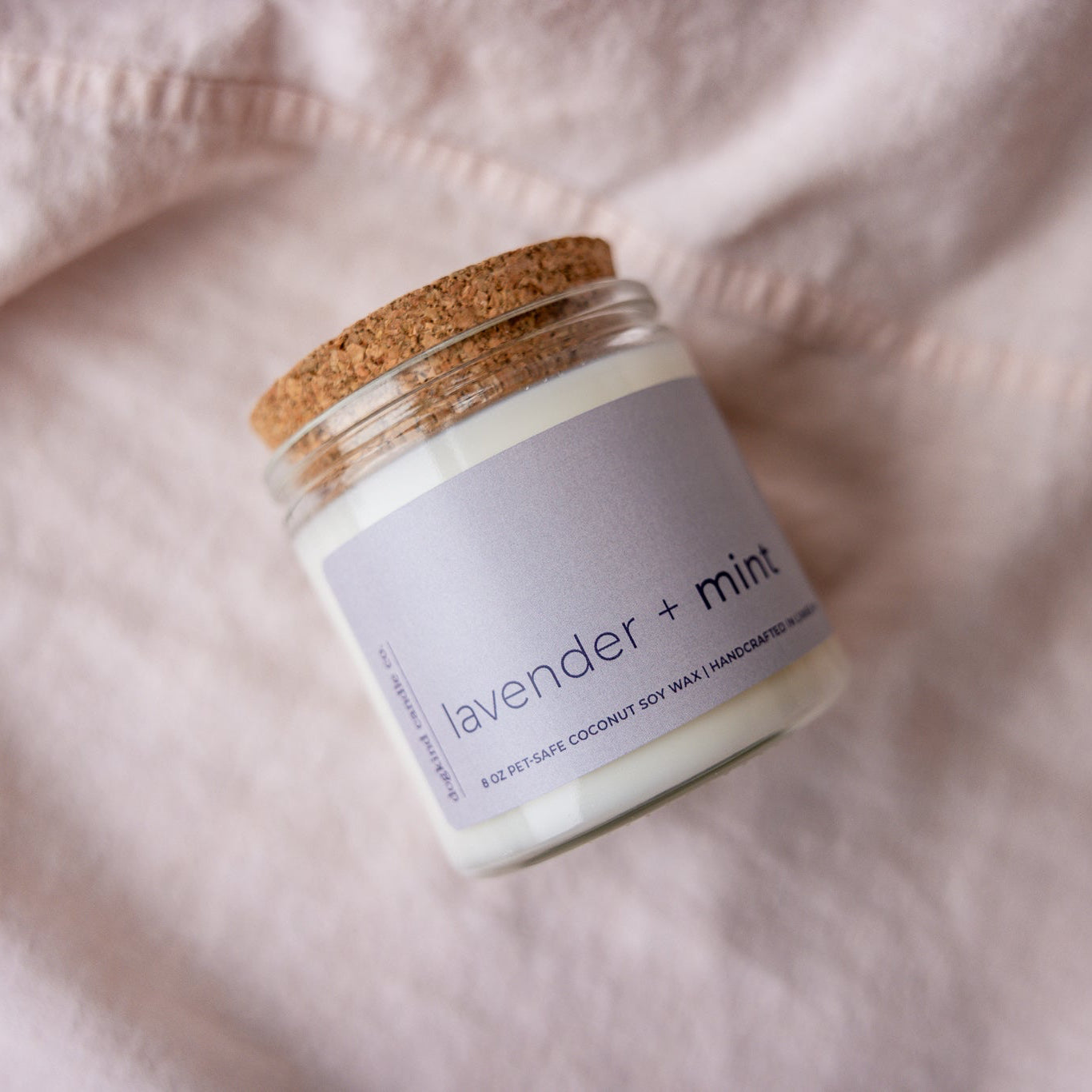 
                  
                    Candle jar labeled 'Lavender + Mint' on a soft pink fabric background
                  
                