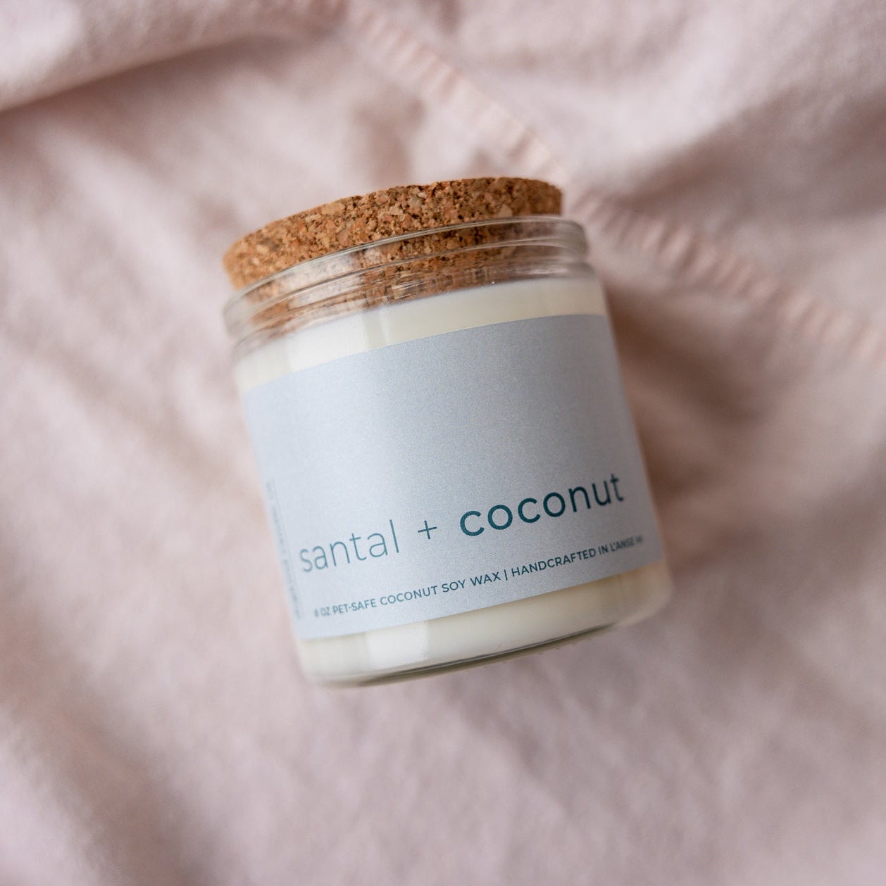 
                  
                    Candle jar labeled 'santal + coconut' on a soft pink fabric background
                  
                