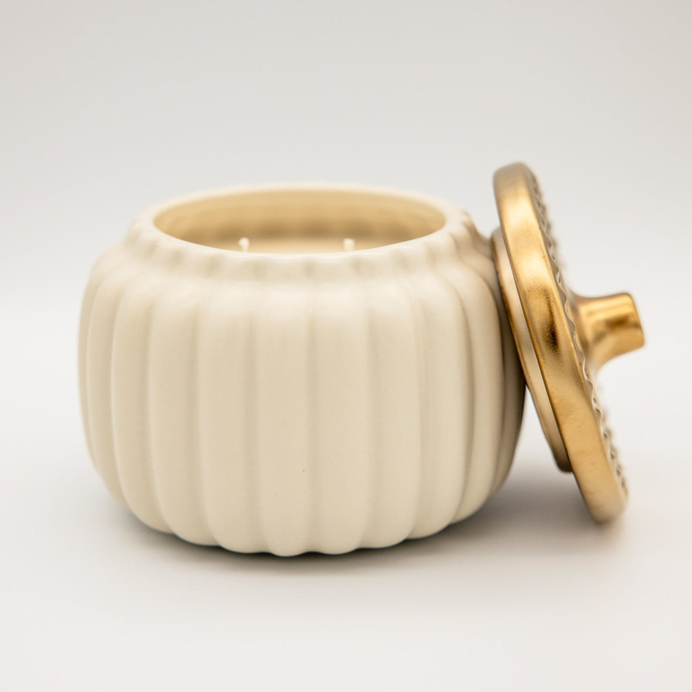 
                  
                    An off-white ribbed ceramic candle jar with its lid propped open, showing two wicks inside. The lid has a metallic gold handle and a textured gold outer rim.
                  
                