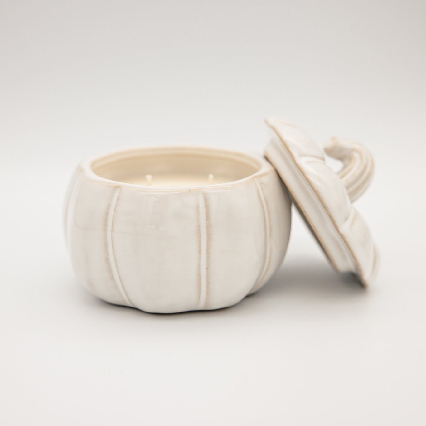 
                  
                    An off-white ceramic pumpkin-shaped candle with its lid leaning against it, set against a seamless light gray background.
                  
                