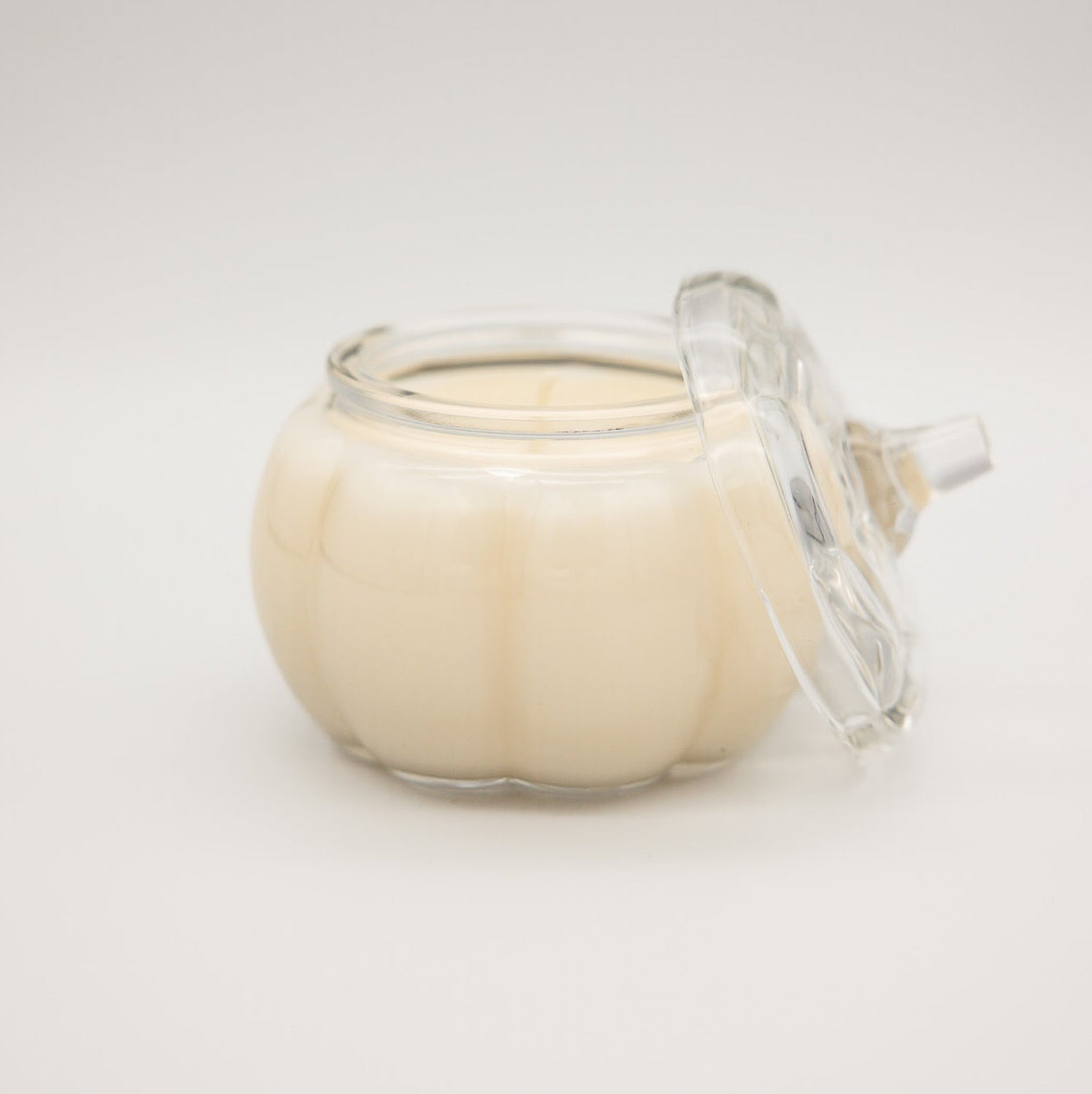 
                  
                    A small clear glass jar candle with a lid leaning against it, filled with creamy off-white wax, photographed on a plain light gray background.
                  
                