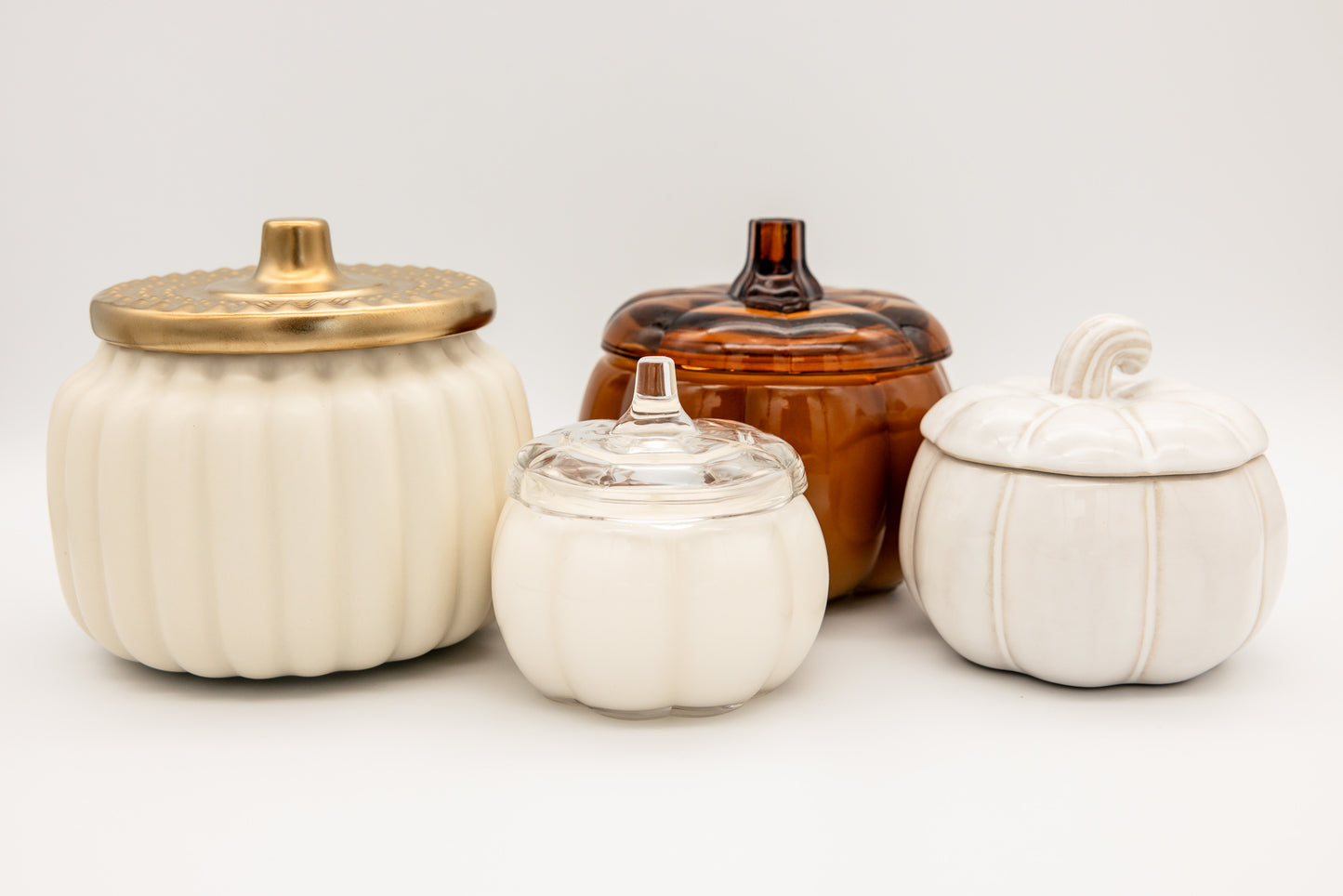 
                  
                    Four decorative pumpkin-shaped lidded jars arranged on a light gray studio background. The set includes two large off-white ribbed jars with metallic gold lids, one medium off-white jar with a clear glass lid, and one amber-brown ribbed jar with a dark stopper.
                  
                