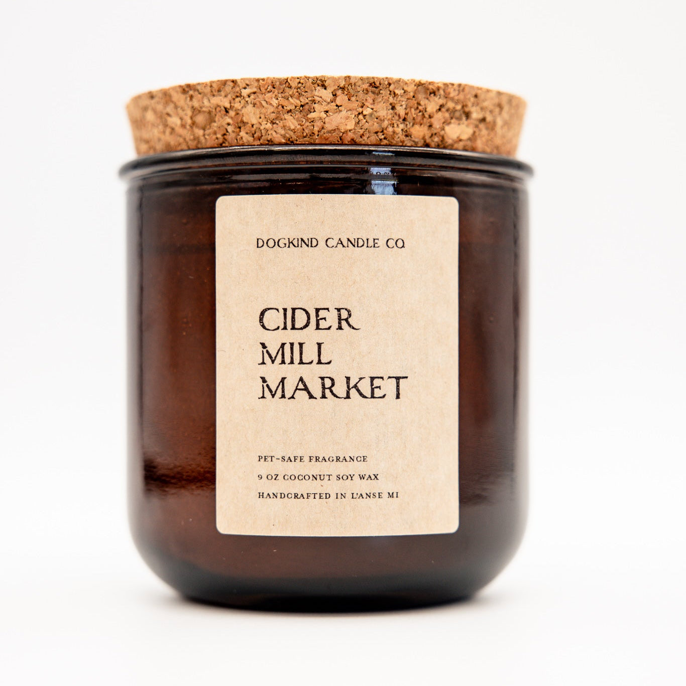 A dark amber glass jar candle with a cork lid and a beige paper label reading 'DOGKIND CANDLE CO' and 'CIDER MILL MARKET' on a white background.