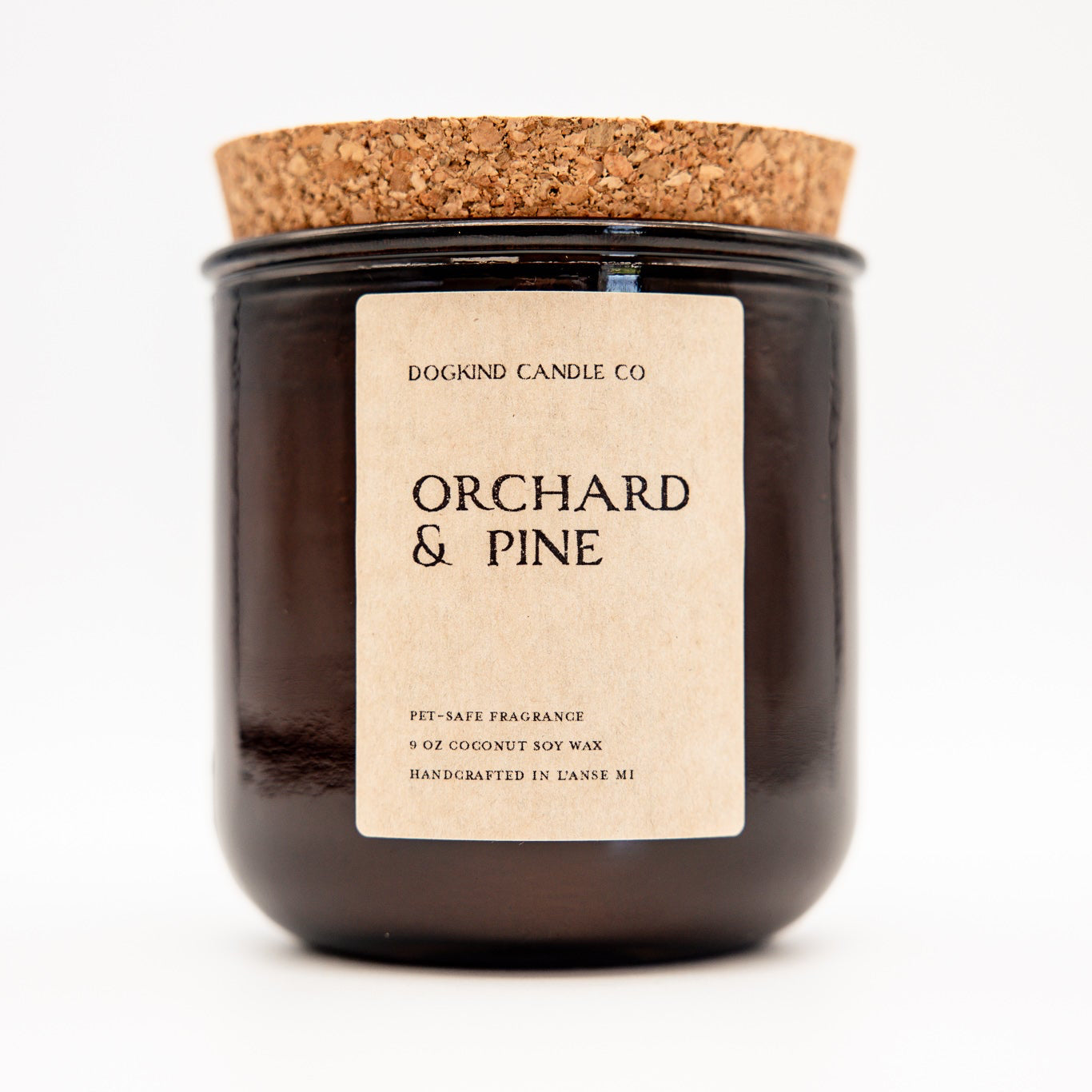 A dark amber glass jar candle with a natural cork lid and a cream paper label reading 'DOGKIND CANDLE CO. ORCHARD & PINE'.