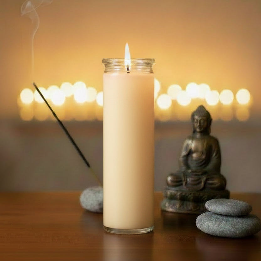 A 7-day cream-colored prayer candle in a clear glass jar stands lit on a wooden surface, with a Buddha statue, smooth stones, and a smoking incense stick in a softly blurred warm-toned background.