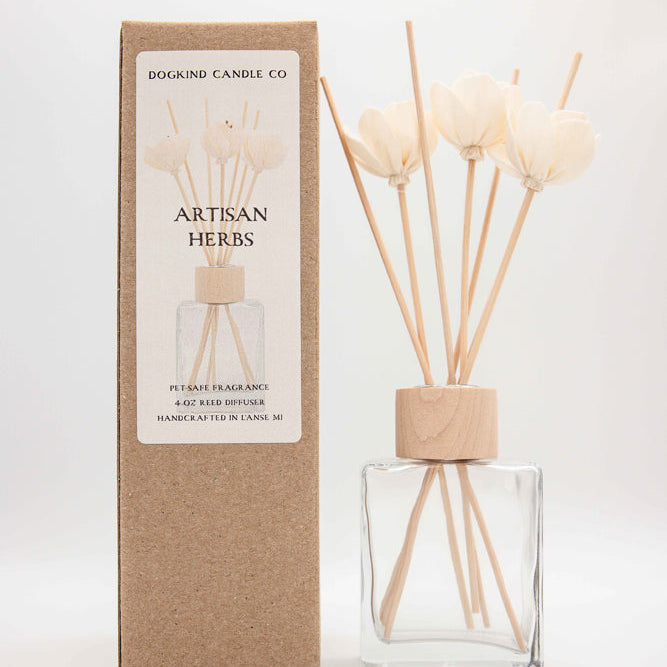 Dogkind Candle Co. Artisan Herbs reed diffuser with a clear glass bottle, natural wood cap, and beige reeds, standing next to its brown kraft paper packaging box on a white background.