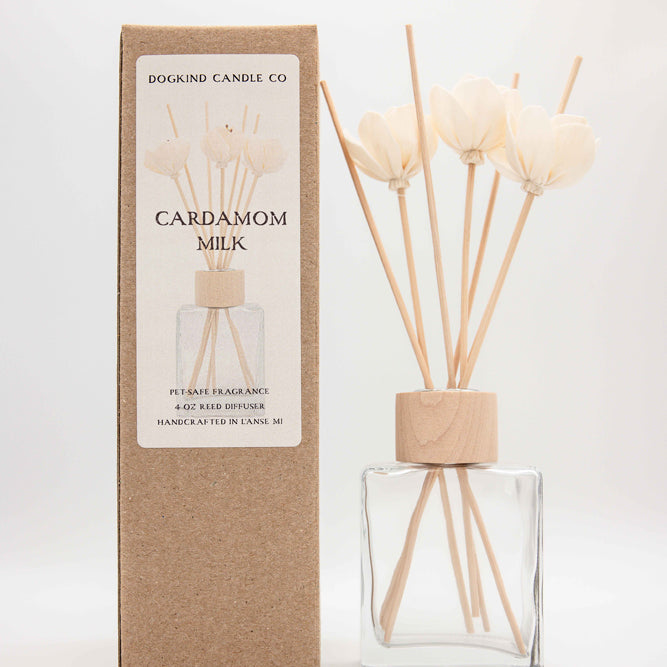 Dogkind Candle Co Cardamom Milk reed diffuser with a clear glass bottle, natural wood cap, and reeds, standing next to its brown kraft paper packaging box on a white background.