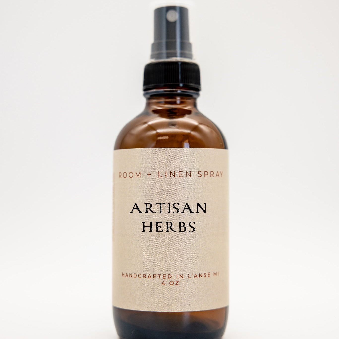 An amber glass spray bottle labeled 'Artisan Herbs' room and linen spray, featuring a black spray nozzle and a cream-colored label, set against a plain white background.