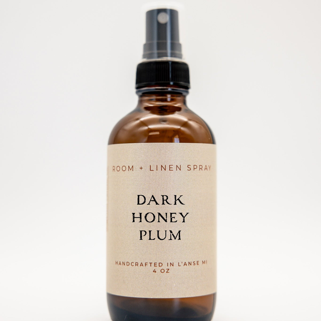 An amber glass spray bottle of Dark Honey Plum Room and Linen Spray with a black spray nozzle and a beige label.