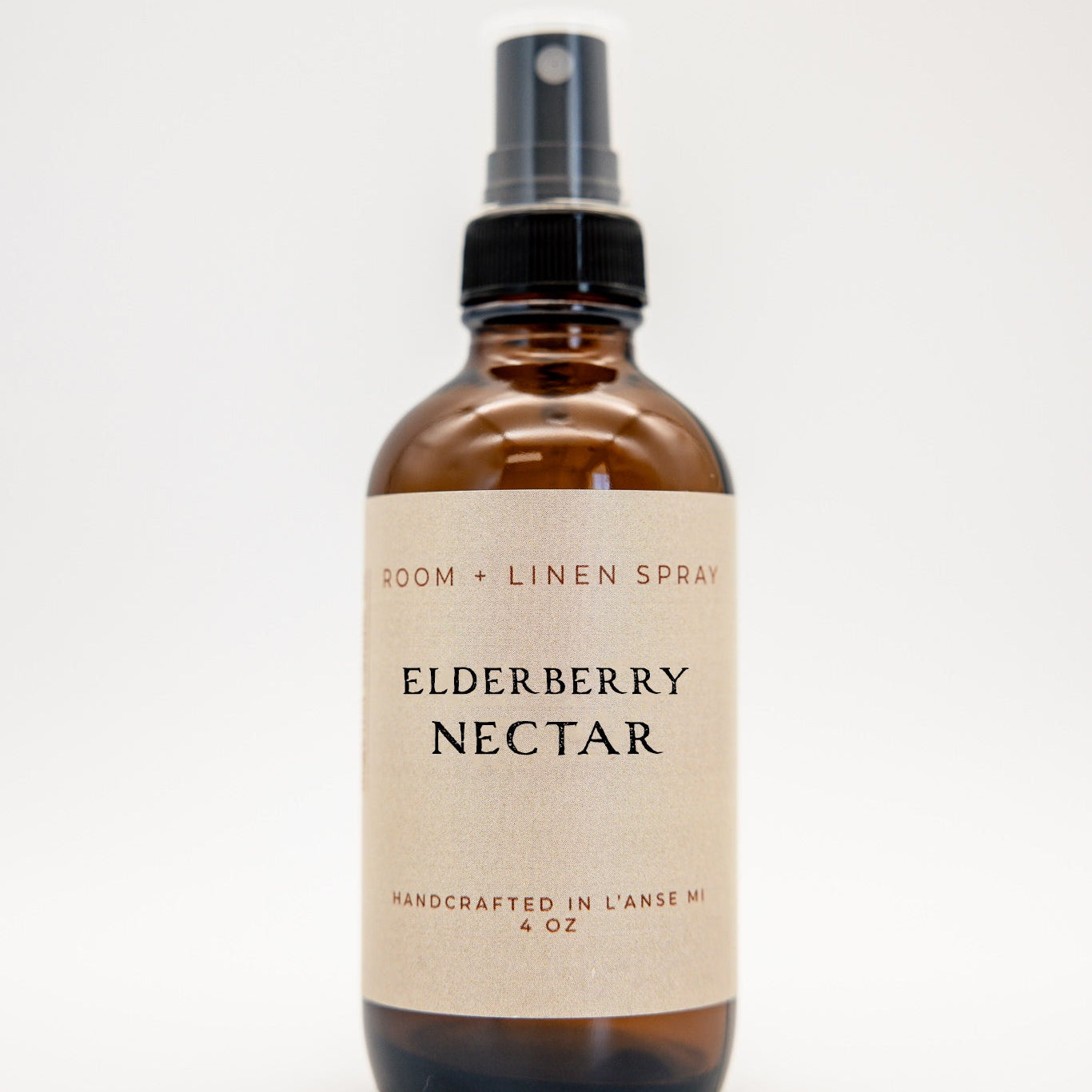 An amber glass spray bottle of Elderberry Nectar room and linen spray with a black spray nozzle and a beige label, set against a plain white background.
