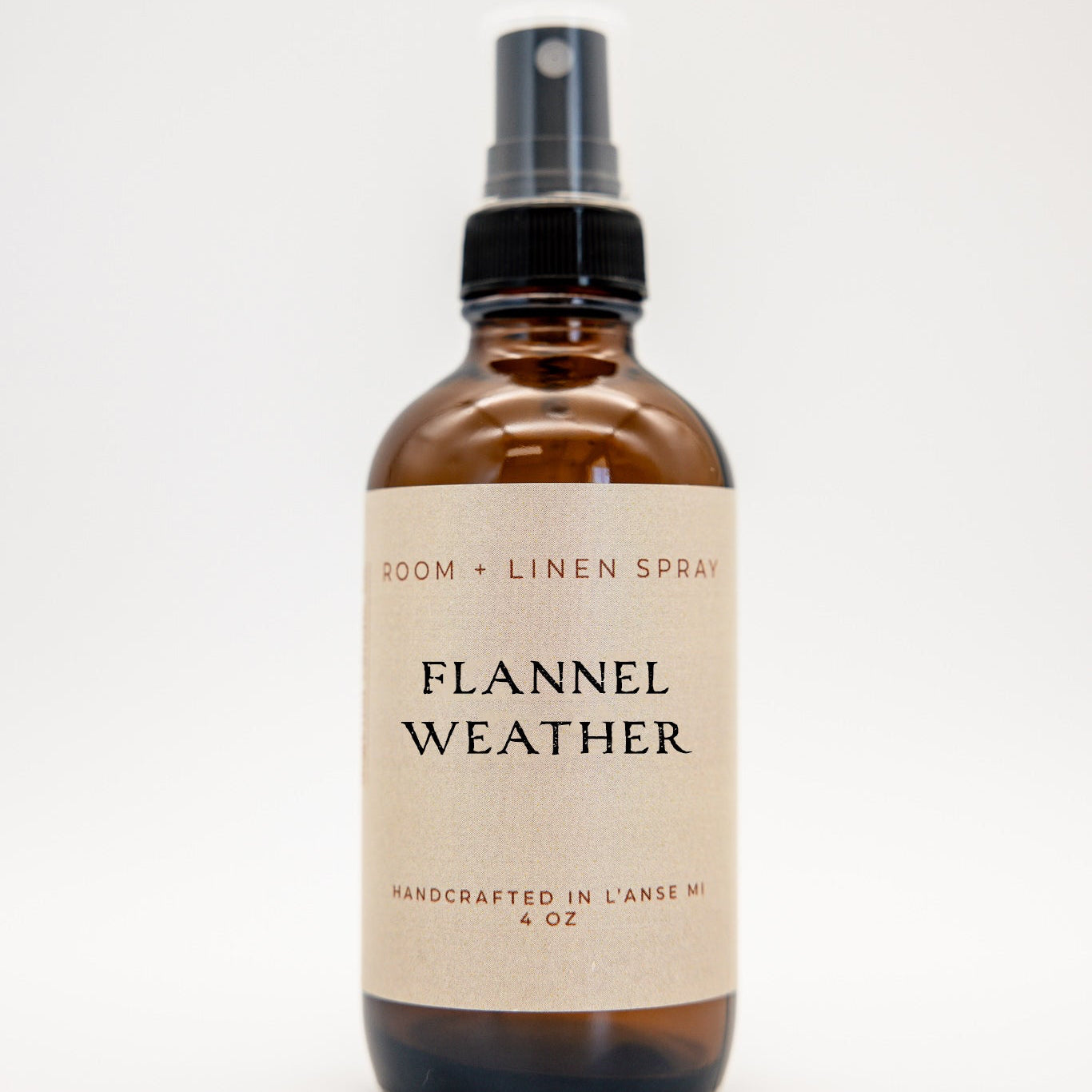 An amber glass spray bottle of 'Flannel Weather' room and linen spray, featuring a cream-colored label and a black spray nozzle, set against a plain white background.