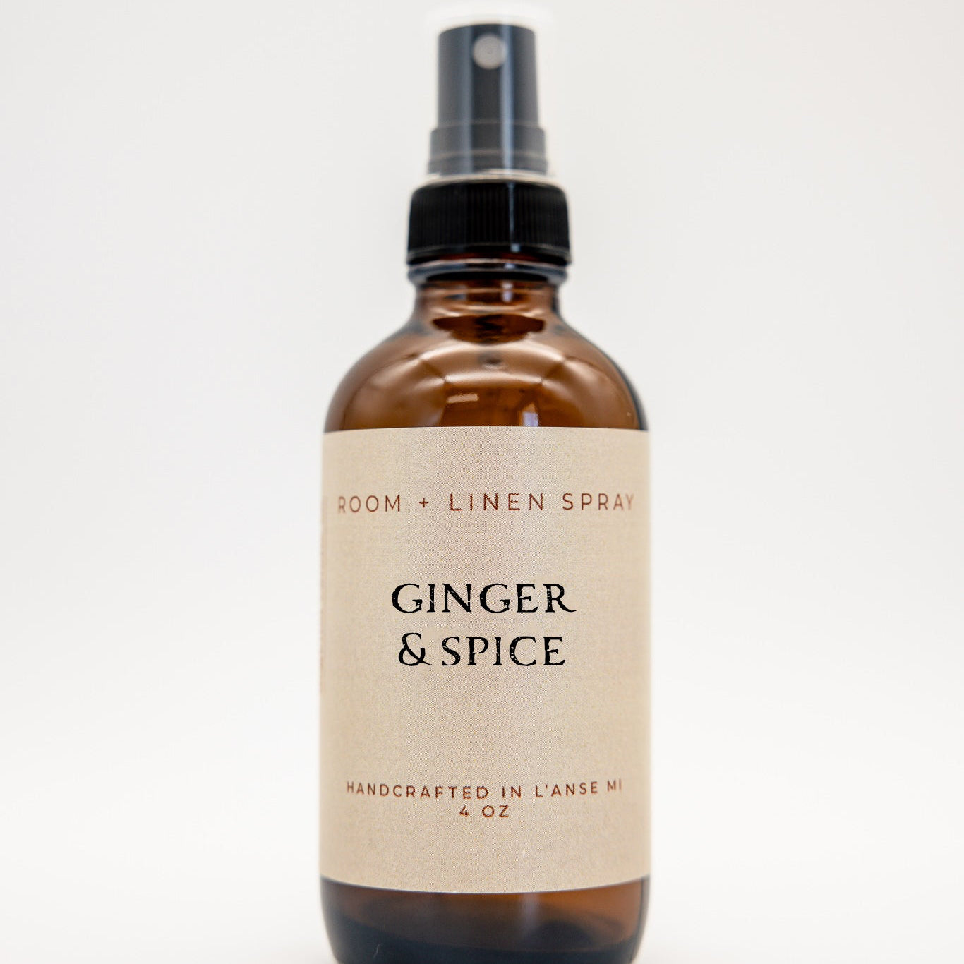 An amber glass spray bottle labeled Ginger & Spice room and linen spray, featuring a black spray nozzle and a cream-colored label, set against a plain white background.