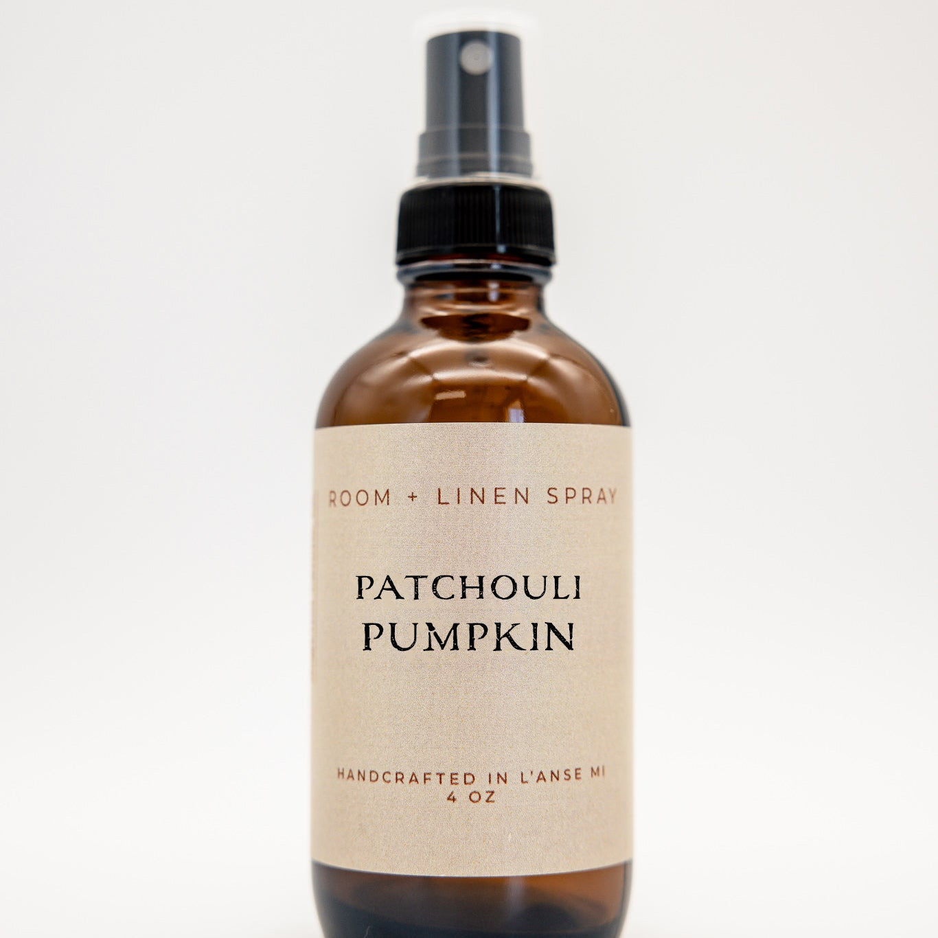 An amber glass spray bottle of Patchouli Pumpkin Room + Linen Spray with a black spray nozzle and a beige label, set against a plain white background.