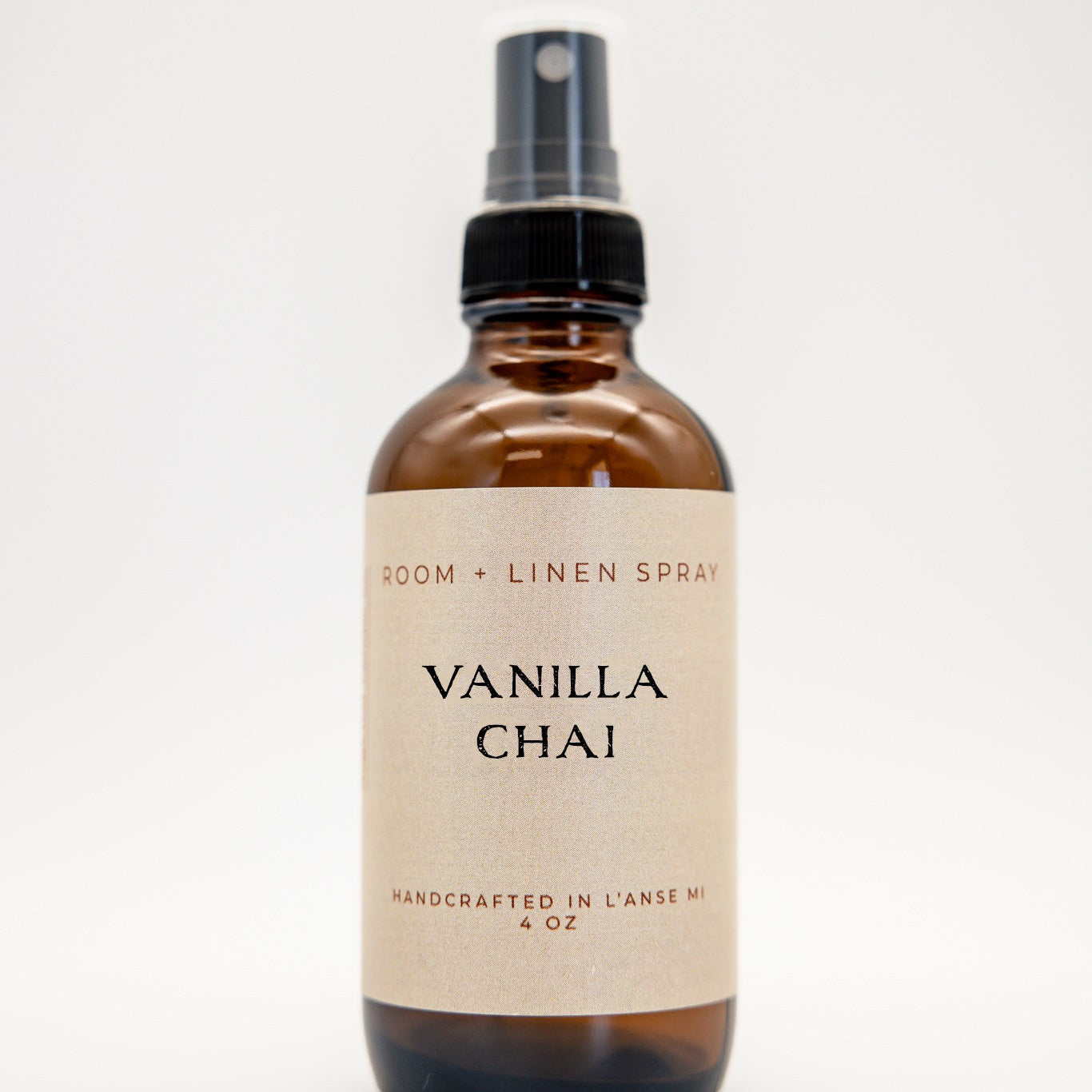 An amber glass spray bottle of Vanilla Chai Room + Linen Spray with a black spray nozzle and a beige label, set against a plain white background.